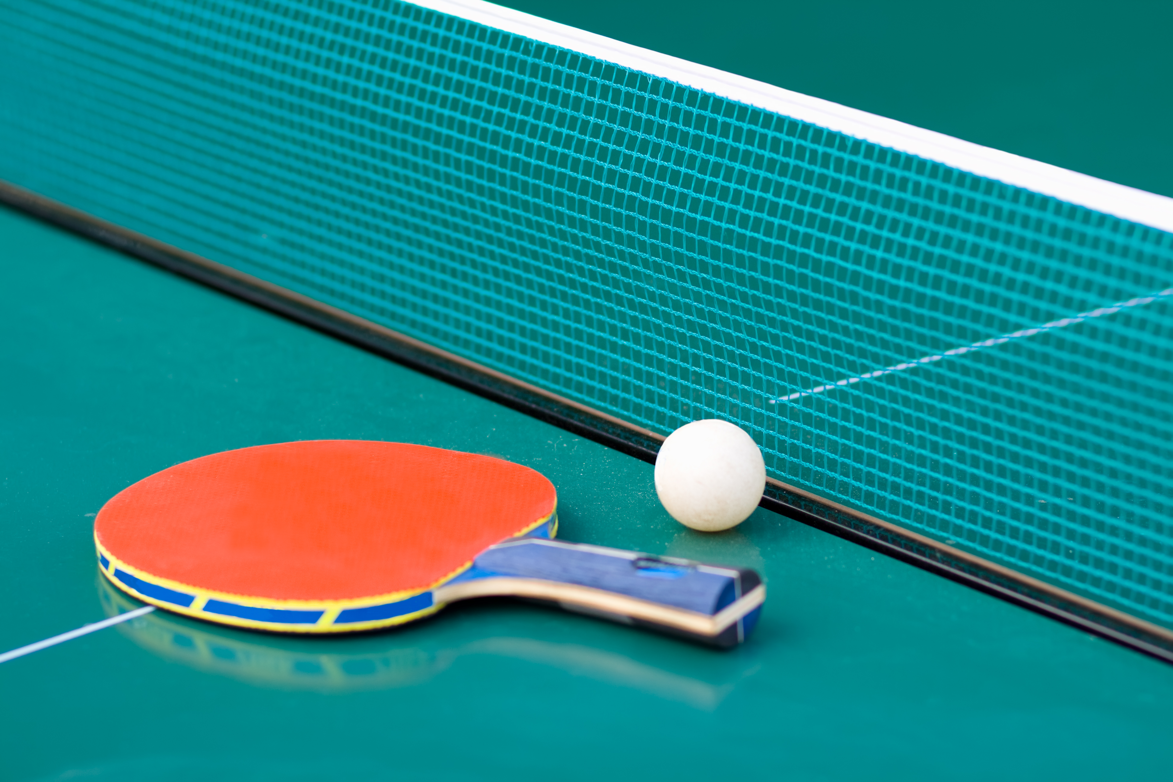 Ping pong bat and ball lying next to a net on top of a green ping pong table.